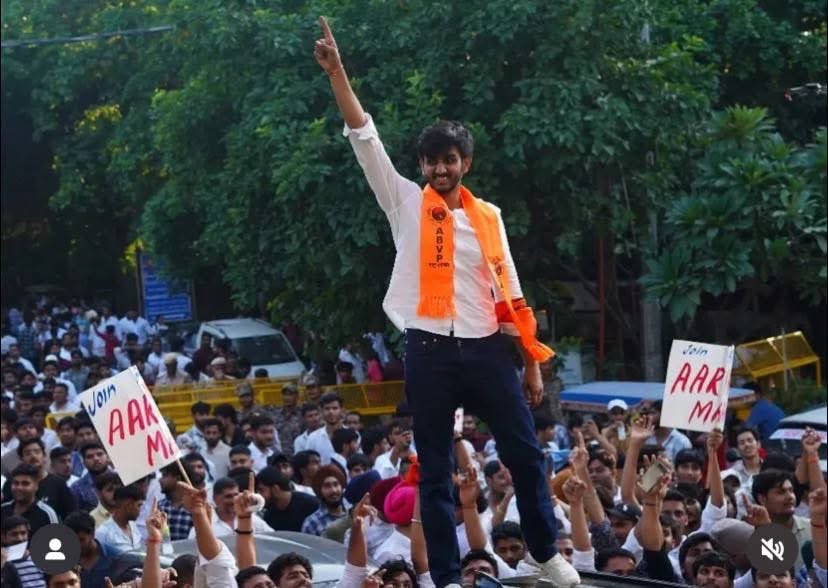 Student leader wearing ABVP scarf raises hand during Delhi University Students Union (DUSU) Election 2025 campaign rally, surrounded by students holding placards.